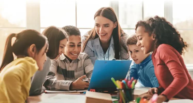 Teacher smiling with a group of students gathered around a laptop in a classroom